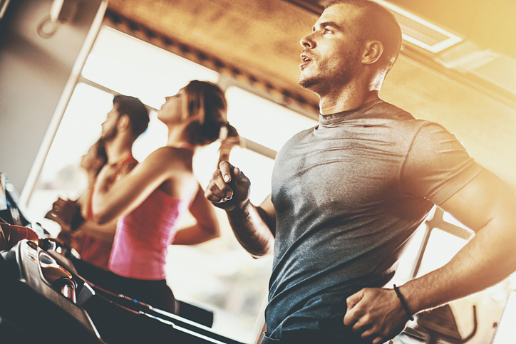 Closeup low angle view of a late 20'sman doing some cardio workout on a treadmill. He's wearing gray sports t-shirt, soaked in sweat.There are three more people next to him, blurry. Back lit, Sun is flaring from the right hand side.