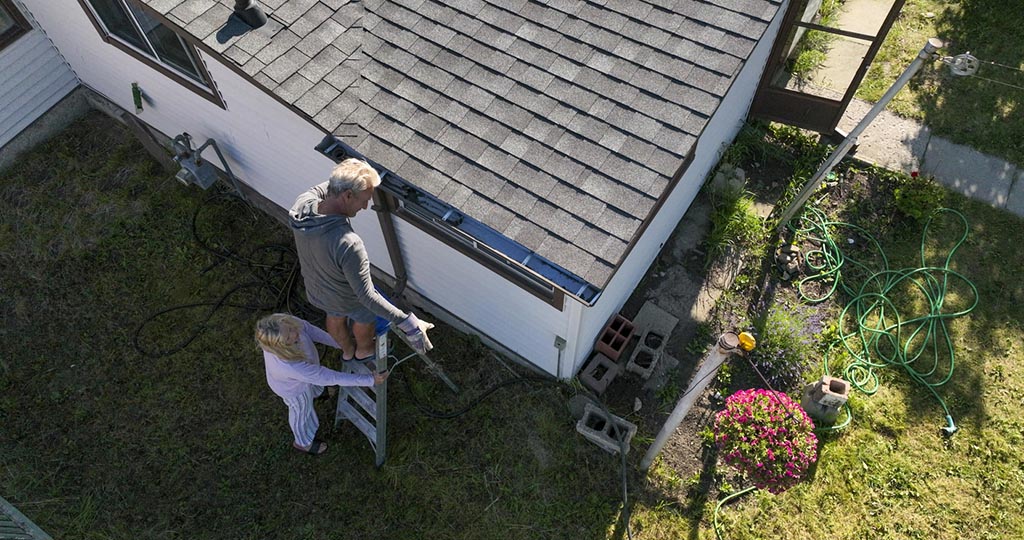 Senior man cleans roof gutter, his partner holds ladder for him